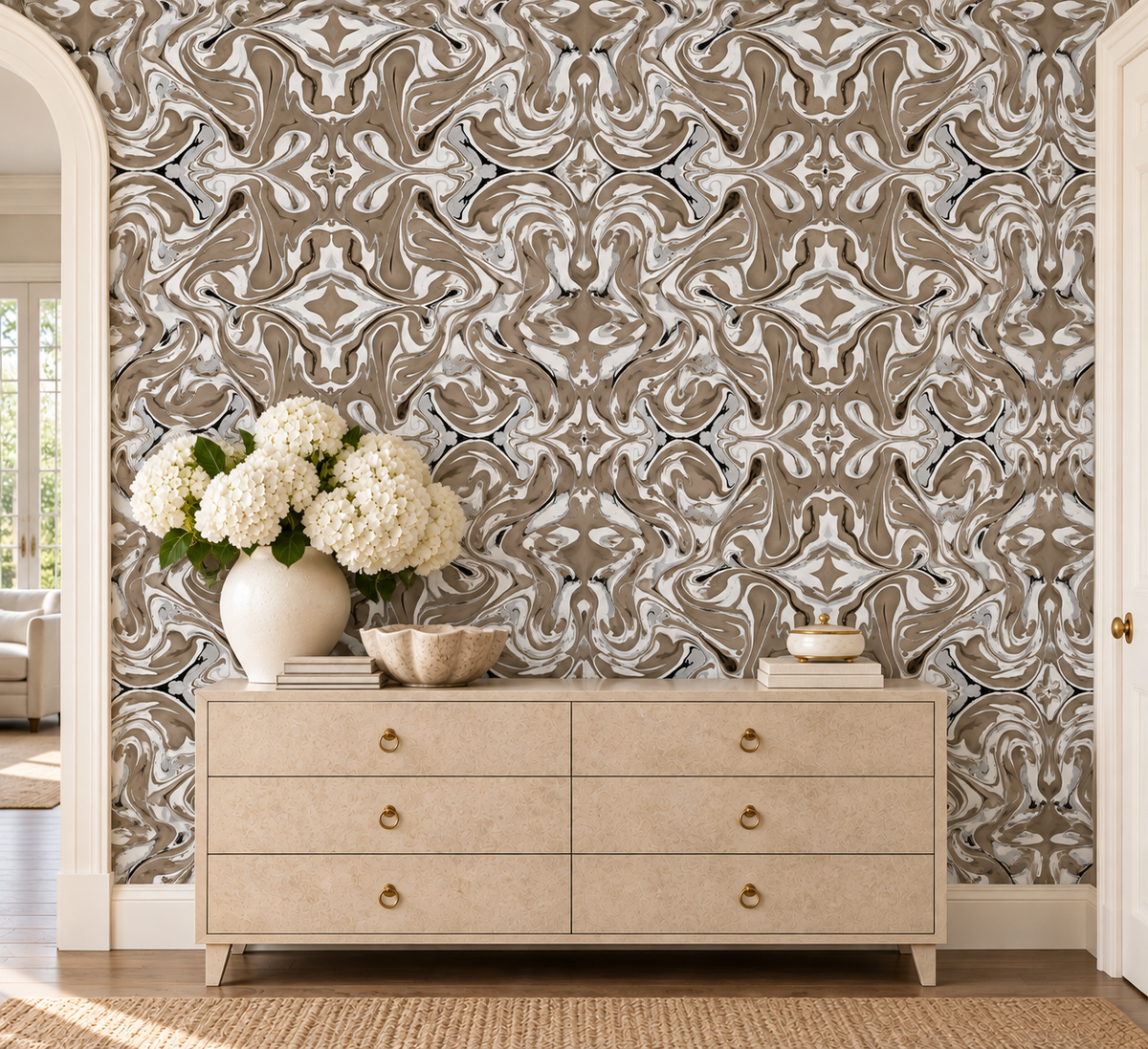 A styled interior entryway featuring a symmetrical wallpaper with an intricate beige, brown, and gray damask pattern. In front of the wallpaper is a light wood dresser with gold knobs, decorated with a white vase of hydrangeas and decorative bowls.